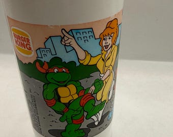 May include: A white plastic cup featuring a Burger King logo and a colorful graphic of a green Teenage Mutant Ninja Turtle character with an orange mask and a person with orange hair in a yellow outfit pointing toward a city skyline background.