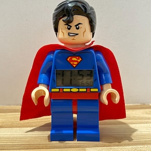 Peut inclure: Un réveil numérique sur le thème de Superman en forme de grande figurine en plastique. Le personnage porte un costume bleu avec une cape rouge, une ceinture jaune et un emblème rouge et jaune sur la poitrine. L'écran affiche 11:53 PM et 6:50.