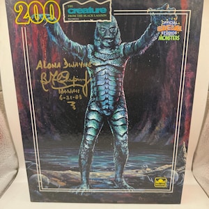 Peut inclure: Boîte vintage avec "The Creature from the Black Lagoon". L'illustration représente la créature les bras levés. La boîte comprend le texte "200", "Official Universal Studios Monsters" et une signature "Aloha Dwayne". La boîte est de Golden.