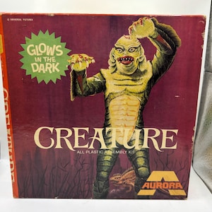 Può includere: Scatola del kit di modelli Aurora vintage con la Creatura della Laguna Nera. La scatola ha uno sfondo rosso scuro con la creatura in verde e giallo. Il testo include "CREATURE" e "GLOWS IN THE DARK". Il logo Aurora è visibile.