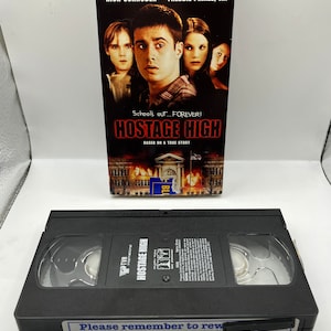 Peut inclure: Une cassette VHS pour le film "Hostage High" avec une image en noir et blanc d'un bâtiment scolaire en feu. Le texte "School's out... FOREVER!" est imprimé sur l'étui. Le film est basé sur une histoire vraie.