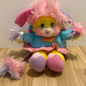 May include: A plush Popples toy featuring a yellow face, pink fur, and blue long-sleeved shirt with a megaphone graphic. The toy has pink and white striped accents on the skirt and feet, and holds two pom-poms in its hands. It has blue eyes.