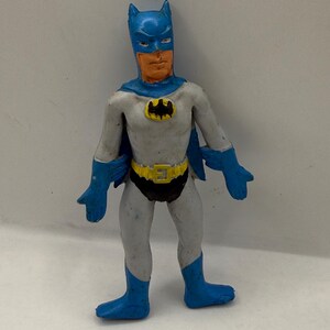 May include: A vintage Batman action figure. The figure is primarily gray and blue, with a yellow bat symbol on the chest. The figure has a blue cowl, cape, gloves, and boots. The belt is yellow with a black buckle.