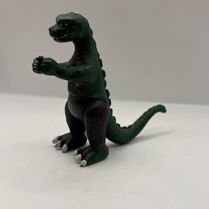 May include: A green Godzilla toy figure with a dark green body and black legs. The toy has a long tail and small white claws. The figure is posed with its arms in front of its chest.