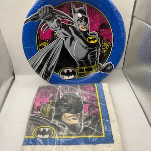 May include: A round blue paper plate featuring a graphic of Batman in a black and grey suit with a yellow emblem, set against a pink and black city skyline. Below is a square pack of paper napkins with a matching Batman graphic and logo.