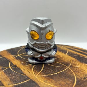 May include: A small, silver figurine with large, orange eyes and a smiling face. The figure has a helmet-like head and is wearing a dark blue and white outfit. It is set on a round, wooden surface with a geometric pattern.