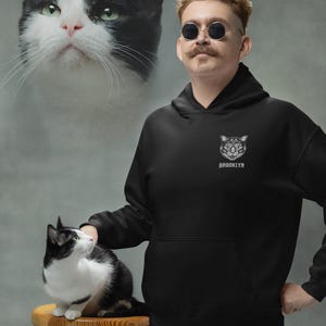 May include: Black hoodie with a cat graphic and the word "BROOKLYN" below it. A person wearing sunglasses and a mustache is standing next to a black and white cat sitting on a wooden stool. A large image of a cat's face is in the background.