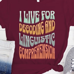 May include: A maroon t-shirt with the text "I live for decoding and linguistic comprehension" in a retro font with colorful accents.