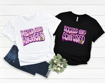 May include: Two t-shirts with a retro font design. One white t-shirt says "Dazed and Engaged" with pink lettering and flowers. The other black t-shirt says "Boozed and Confused" with pink lettering and flowers.
