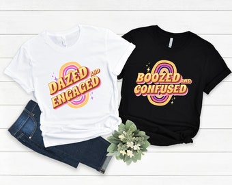 May include: Two t-shirts, one white and one black, with a retro rainbow design and text. The white shirt says "Dazed and Engaged" and the black shirt says "Boozed and Confused".