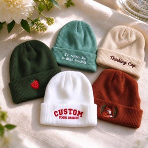 Op de afbeelding: Vijf gebreide mutsen in verschillende kleuren: een donkergroene met een aardbei, een groenblauwe met de tekst "I'd rather be in MAS Foodhall", een crèmekleurige met "Thinking Cap", een witte met "CUSTOM YOUR DESIGN" en een roestkleurige met een vos.