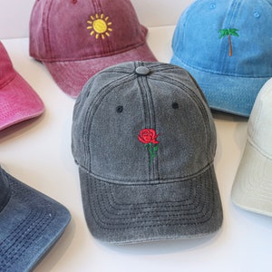 Op de afbeelding: Zes verschillende gekleurde denim baseballpetten met geborduurde ontwerpen. De petten zijn in een cirkel op een wit oppervlak gerangschikt. De ontwerpen omvatten een vlinder, een zon, een palmboom, een roos, een regenboog en een golf.