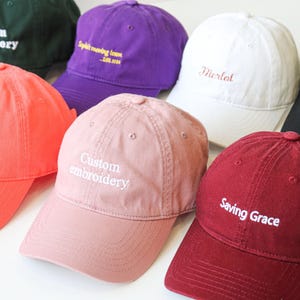 Könnte beinhalten: Eine Sammlung von zehn Baseballkappen in verschiedenen Farben mit unterschiedlichen gestickten Texten. Der Text umfasst "Custom Embroidery", "It's Go Baby", "Merlot", "4.6.7.II", "Saving Grace" und "Spilt Morning Love - Los Altos".