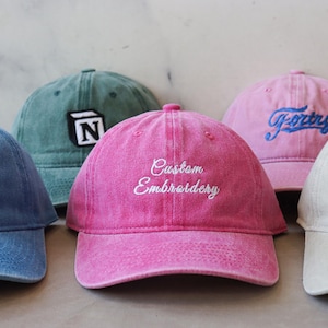 Pode incluir: Cinco bonés de beisebol de diferentes cores com texto bordado. Os bonés são azuis, verdes, rosa, brancos e bege. O texto nos bonés diz "Down South", "N", "Custom Embroidery", "Goofy" e "Romantic".