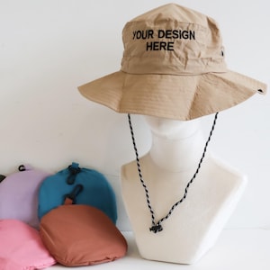 May include: A tan sun hat with a wide brim and the text "YOUR DESIGN HERE" printed on the crown. The hat is displayed on a mannequin head with a black chin strap. Several colorful, folded pouches in shades of green, pink, purple, teal, and brown are in front.