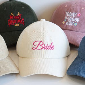 Könnte beinhalten: Eine Sammlung von Cord-Baseballkappen in verschiedenen Farben, jede mit einzigartigem Text bestickt. Die Kappen enthalten Sätze wie "Bride", "Dad EST. 2025", "Merry Christmas" und "Today is a good day". Eine Kappe ist anpassbar.