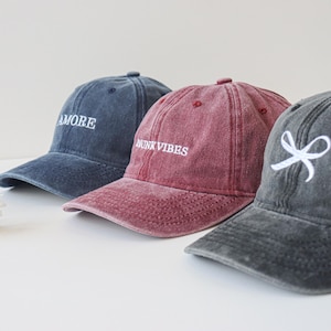 Könnte beinhalten: Drei Baseballkappen aus Denim mit verschiedenen Stickereien. Die erste Kappe ist blau mit dem Wort "AMORE" in weiß gestickt. Die zweite Kappe ist burgunderrot mit den Worten "DRINK VIBES" in weiß gestickt. Die dritte Kappe ist dunkelgrau mit einer weißen Schleife, die auf der Vorderseite gestickt ist.