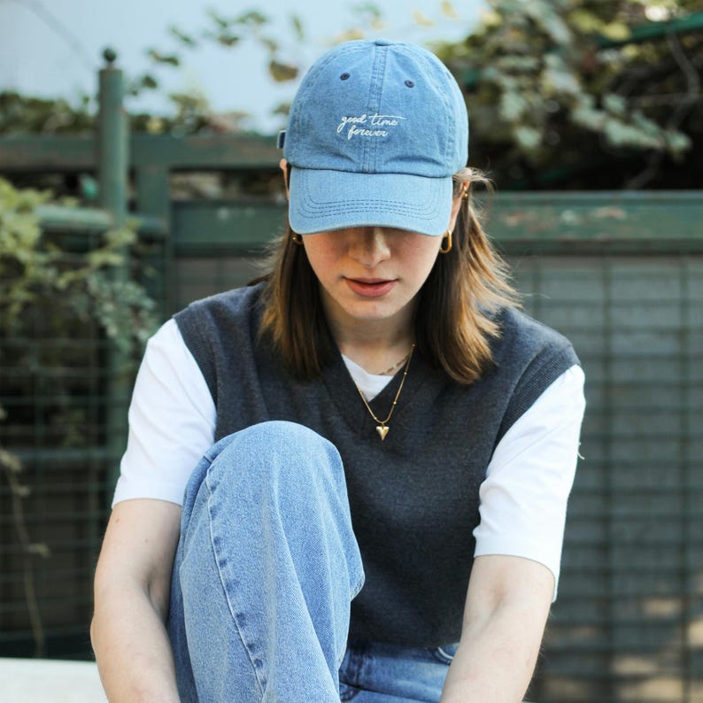 Pu&ograve; includere: Una persona indossa un berretto di jeans blu con il testo "good time forever" ricamato sul davanti. Indossa una maglietta bianca e un gilet grigio. La persona indossa jeans blu.