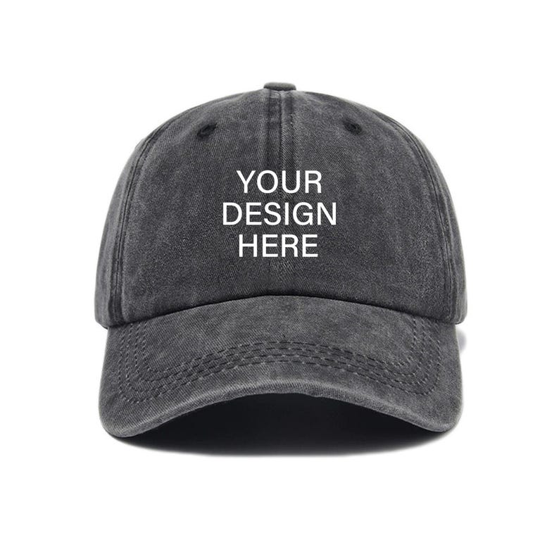 Pu&ograve; includere: Un cappello da baseball in cotone nero, usurato, con il testo "YOUR DESIGN HERE" in bianco.