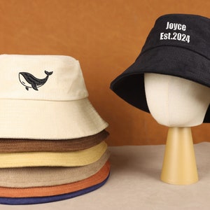 Op de afbeelding: Een stapel van zes bucket hats in verschillende kleuren, waaronder beige, bruin, geel en oranje. De bovenste hoed heeft een zwarte walvis geborduurd. Een zwarte bucket hat met de tekst "Joyce Est. 2024" wordt getoond op een standaard.