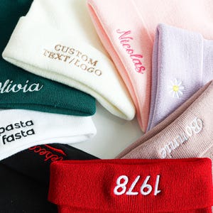 Op de afbeelding: Een verzameling gebreide mutsen in verschillende kleuren, waaronder wit, groen, roze, paars en rood. Sommige mutsen zijn geborduurd met namen, tekst of logo's. Eén muts heeft de tekst "eat pasta run fasta", een andere het nummer "1978".