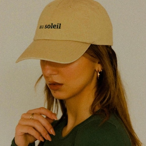 Pu&ograve; includere: Un cappello da baseball beige con il testo "au soleil" ricamato sul davanti.