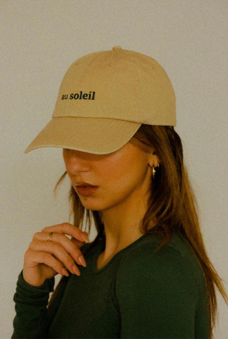 Pu&ograve; includere: Un cappello da baseball beige con il testo "au soleil" ricamato sul davanti.