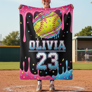 May include: A black softball blanket with a colorful design. The blanket features a yellow softball with rainbow stripes and pink, blue, and yellow dripping accents. The name "OLIVIA" and the number "23" are displayed in a blue and white font.