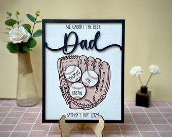 May include: A white framed sign with the text "We caught the best Dad" in black lettering. A baseball glove with three baseballs inside is illustrated. The baseballs have the names "Thomas", "Jake", and "Dustin" written on them. The sign also says "Father's Day 2024".