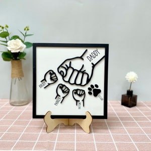 May include: Black and white wood wall art with a design of five fists, one larger than the others, with the names "Daddy", "Brody", "Parker", "Patton", and "Ozark" written below each fist. The art is on a black frame and a wooden stand.