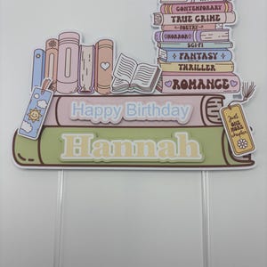 May include: A birthday cake topper with a book theme. It features a stack of books with genre labels, the words "Happy Birthday Hannah", a bookmark, and a tag that says "Just one more chapter."