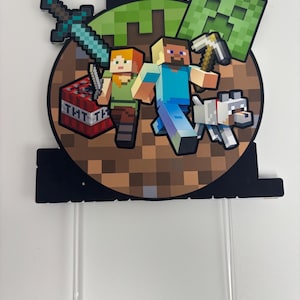 May include: A Minecraft cake topper with pixelated characters Steve and Alex, a white dog, a creeper, a sword, and a TNT block. The topper is attached to clear plastic sticks for easy insertion into a cake.