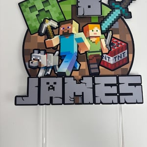 May include: A Minecraft cake topper with pixelated characters, a sword, and TNT. The name "JAMES" is displayed in grey block letters. The topper includes a green creeper, a wolf, and two figures with tools.