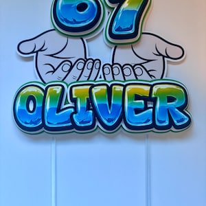 May include: A cake topper with the number "67" in blue, yellow, and green bubble letters, above the name "OLIVER" in similar colors. The design includes cartoon hands and is mounted on clear sticks.