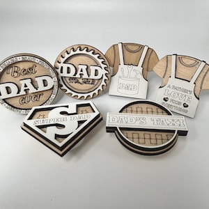 May include: A selection of wooden decorations for Father's Day. The designs feature the phrases "Best Dad Ever", "Dad Mr. Fix It", "Super Dad", and "Dad's Taxi". Other designs include a shirt and apron with the words "Dad" and "A Father's Love Fixes Everything".