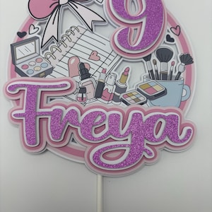 May include: A pink and white cake topper with the name "Freya" in glittery purple, the number "9", and makeup-themed illustrations. Includes a bow, hearts, and a notepad. The topper is attached to a white stick.