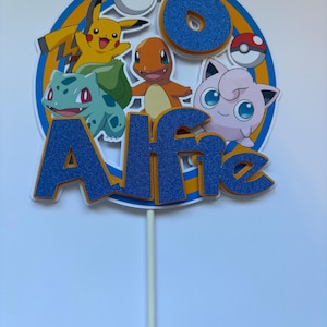 May include: A circular cake topper featuring the name Alfie in blue glittery letters and the number 6 in blue glitter. The design includes illustrations of Pikachu, Charmander, Bulbasaur, and Jigglypuff, along with two Poké Balls on a white stick.