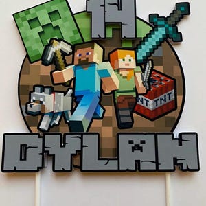 May include: A Minecraft cake topper with pixelated characters, a wolf, and a TNT block. The design includes the number "14" and the name "DYLAN" in grey block letters. The topper is attached to white sticks.