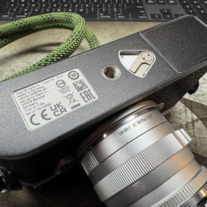 May include: A black digital camera with a silver lens. The camera has a black leather strap and a silver metal plate with the text "Digital Camera - Type No.: 2416" and "Made in Germany" printed on it.