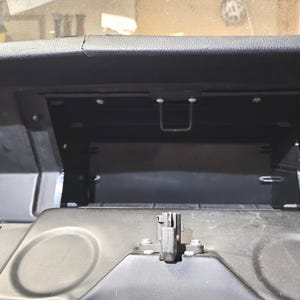 May include: An open black automotive glove box compartment featuring a textured plastic interior, a central latch mechanism, and two circular indentations on the exterior door panel, set within a dark dashboard assembly with visible mounting screws.