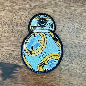 May include: A blue and yellow embroidered patch of a Star Wars character, BB-8, on a wood background.