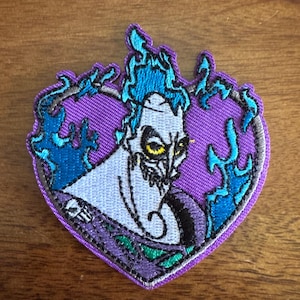 May include: A heart-shaped patch featuring a cartoon character with blue flames and a purple background. The character has pale skin, a black and purple outfit, and yellow eyes. The patch is embroidered with various colors.