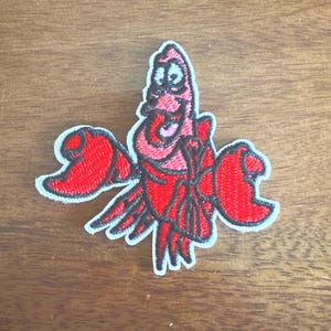May include: A red and pink embroidered patch featuring a cartoon crab with a smiling expression. The crab has large red claws and is outlined in black. The patch has a white border and is likely for clothing or accessories.