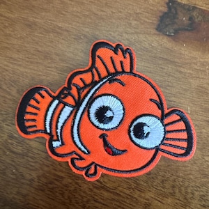 May include: Embroidered patch featuring a cartoon clownfish in orange, white, and black. The fish has large, expressive eyes and a smiling mouth. The patch is likely for clothing or accessories.