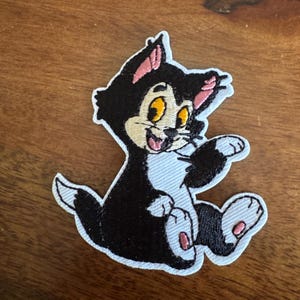 May include: A black and white embroidered patch featuring a cartoon cat with yellow eyes and pink accents. The cat is sitting with its paws outstretched, smiling. The patch has a white border.