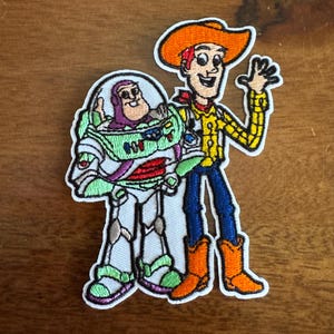 May include: Embroidered patch featuring cartoon characters Buzz Lightyear and Woody from Toy Story. Buzz is in a white and green spacesuit, and Woody is wearing a cowboy hat, yellow shirt, blue pants, and orange boots. The patch has a white border.