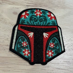 May include: Embroidered patch featuring a stylized helmet design. The helmet is primarily black with red and teal accents. Floral patterns in red and white adorn the teal sections, creating a unique decorative element.