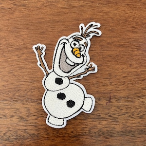 May include: Embroidered patch featuring Olaf from Frozen. The patch depicts Olaf with a smiling expression, raised arms, and a carrot nose. The patch is white with black, brown, and orange details. The patch is approximately 3 inches tall.