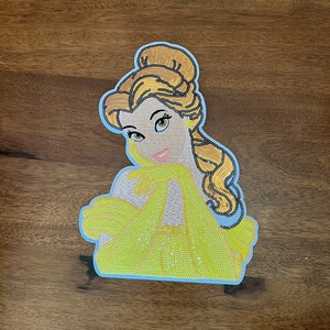 May include: A decorative patch featuring a stylized illustration of a character with brown hair styled in an updo, wearing a bright yellow off-the-shoulder dress and matching yellow gloves, with a light blue border outlining the entire shape.