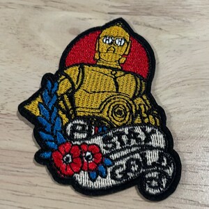 May include: Embroidered patch featuring C-3PO from Star Wars, in gold and black, set against a red background. The patch includes the text "Stay Gold" in a banner with floral and leaf accents.